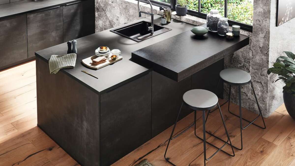 Stilvolle Küche mit schwarzer Betonarbeitsfläche, integriertem Spülbecken, zwei Hockern und großen Fenstern mit Blick auf Grünpflanzen.